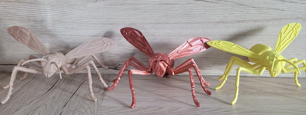 Trois insectes articulés imprimés en 3D côte �à côte, en beige, cuivre et jaune, posés sur un plan de bois clair